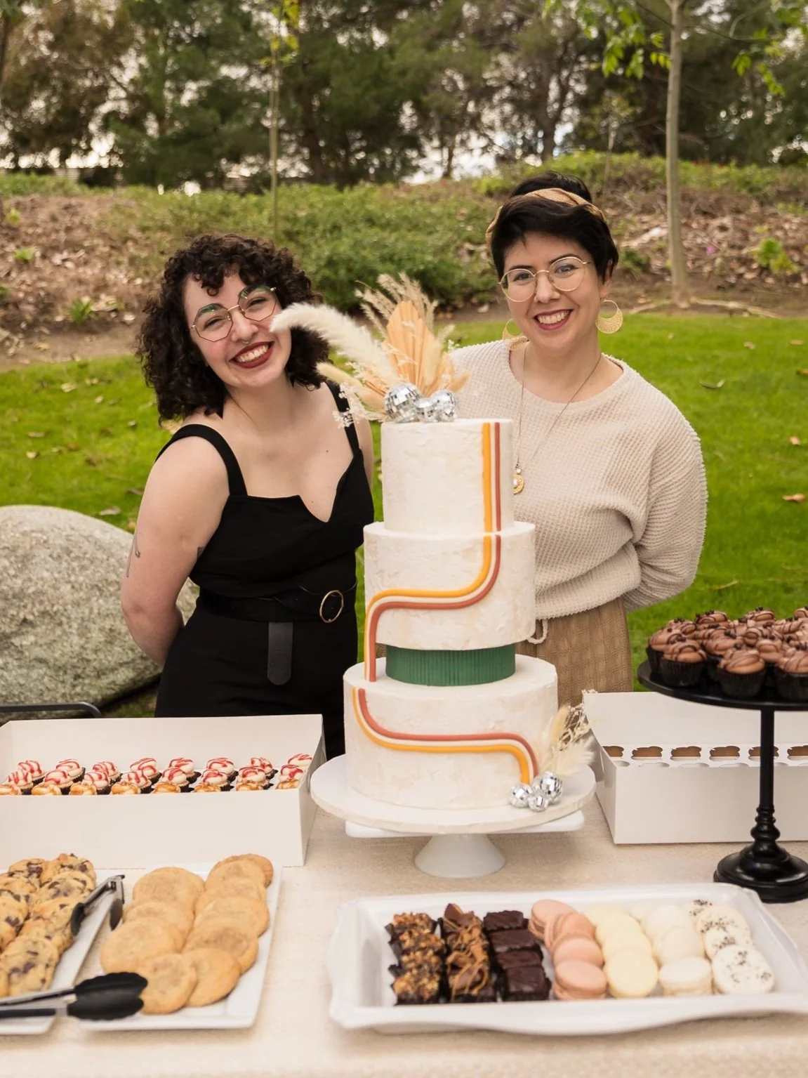 Yanett Ortiz and Debbie Andrade smiling behind a dessert table with a three-tier retro cake, cookies, brownies, macarons and cupcakes